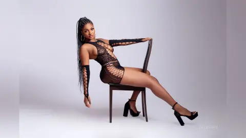 IvyBerry poses on a chair wearing a black fishnet bodysuit and black high heels, showing off her long braids and bare legs.