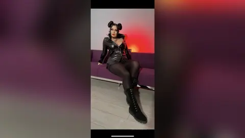 Woman in edgy black leather jacket and boots sitting on a purple sofa in a moody studio setting.