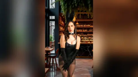Woman in black bunny ears and bodysuit posing in a moody, upscale restaurant setting.