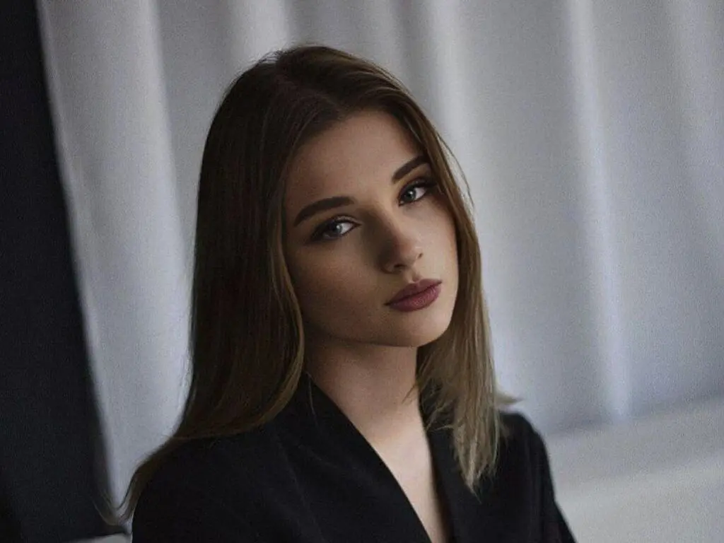 Portrait of a young woman with long brown hair wearing a black outfit in a soft, moody indoor setting.