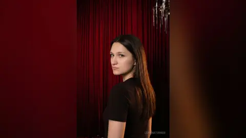 Young woman in a black top posing against elegant red velvet curtains in a moody, cinematic setting.