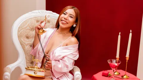Smiling woman in a pink satin robe sitting in a white vintage chair against a red background with candles.