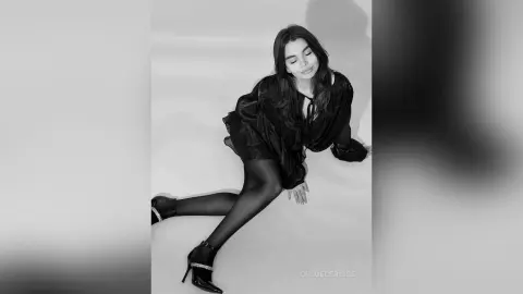 Elegant woman in a black silk dress and high heels sitting against a white background in a moody black and white portrait.