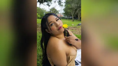 Smiling young woman with long dark hair holding a yellow flower in a lush green outdoor garden setting.