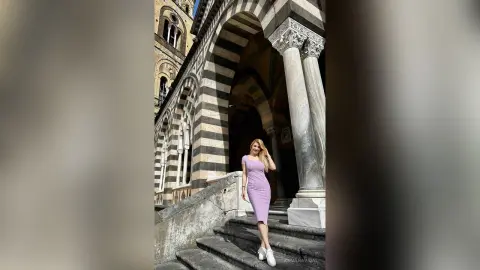 Smiling woman in a lavender midi dress standing on stone stairs of a historic striped cathedral.