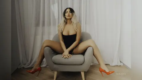 IzieAiko sits in a grey chair wearing a black bodysuit and red high heels, spreading her legs wide with her hands resting between her thighs.