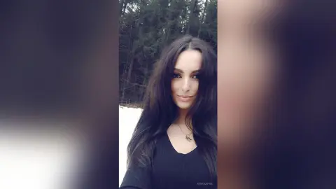AishaApril poses outdoors with long dark hair, wearing a black v-neck top against a background of dark trees and snow.