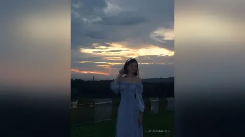Young woman in a flowing light blue dress standing under a dramatic sunset sky in a dreamy outdoor setting.
