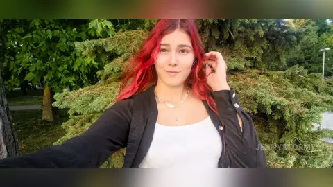 Smiling young woman with red hair wearing a black jacket and white top in a lush green garden setting.