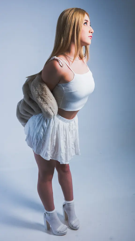 Sophiie_Collins — Sophiie_Collins stands in profile wearing a white tank top and white skirt with a tan fur shrug.