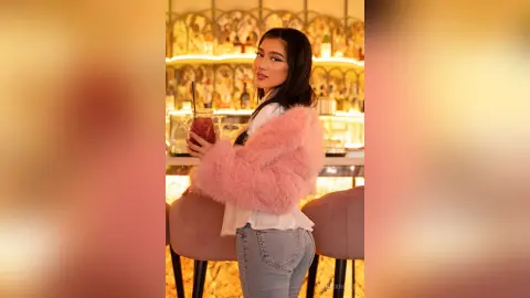 Young woman in a pink faux fur jacket holding a cocktail in a warm, glowing upscale bar setting.