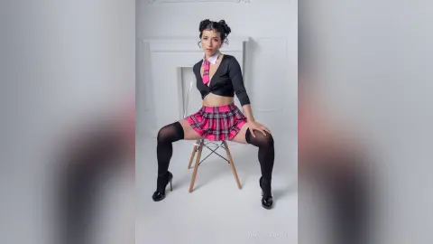 Young woman in a black crop top, pink plaid skirt, and tie sitting on a chair in a bright minimalist studio.