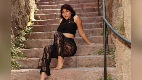 Brunette woman in black lace crop top and sheer trousers sitting on stone stairs in a warm, natural setting.