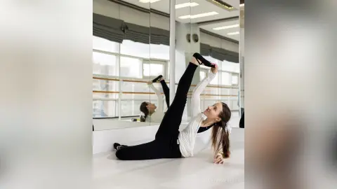 Flexible woman in white long sleeve shirt and black leggings stretching on floor in bright dance studio.