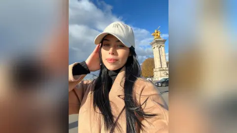 Stylish woman in a tan coat and white cap smiling near the golden statue at Place de la Concorde, Paris.