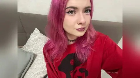 Young woman with pink hair and winged eyeliner wearing a red graphic t-shirt in a cozy bedroom setting.