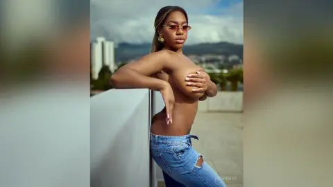 Topless Black woman in denim jeans posing outdoors against a city skyline background.