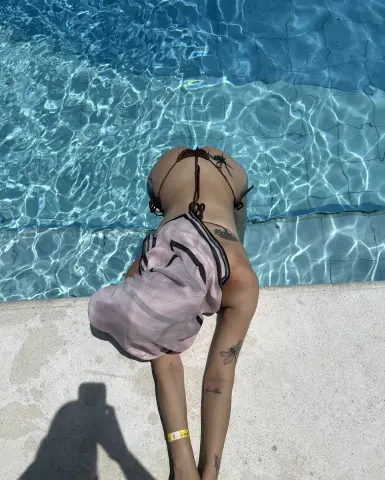 Woman in a pink cover-up leaning over a bright blue swimming pool on a sunny summer day.