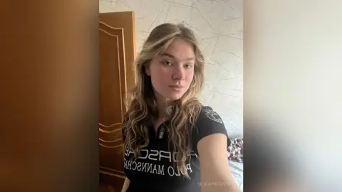 SilviaPrespertys is a blonde woman wearing a black t-shirt, posing for a selfie in her bedroom.