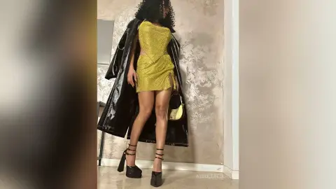 NicoleBella stands in a black shiny trench coat wearing a short, tight yellow glitter dress and black strappy heels.