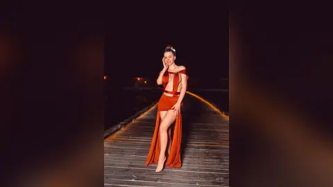 Elie poses on a wooden pier at night wearing a long, burnt-orange dress with a deep plunging neckline and a high slit showing her leg.