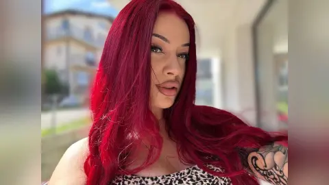 AriaVex poses with her long red hair flowing over her shoulders, wearing a black and white leopard print top.