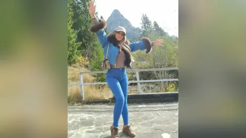Cheerful woman in blue denim outfit and fur-trimmed jacket posing joyfully against a scenic mountain landscape.