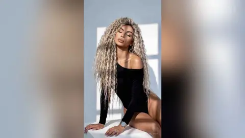 Glamorous woman with long wavy blonde hair wearing a black long-sleeve bodysuit in a bright minimalist studio.
