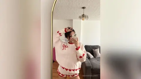Young woman in a white and red heart pattern sweater posing in a mirror for a cozy aesthetic selfie.