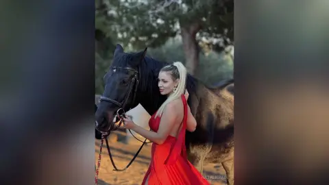 NatalieRain poses outdoors in a long red dress while standing next to a black horse, with her blonde hair pulled back in a ponytail.