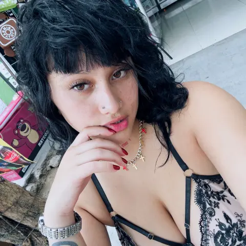 Young woman with wavy black hair and nose ring wearing a black lace camisole in a bright outdoor setting.