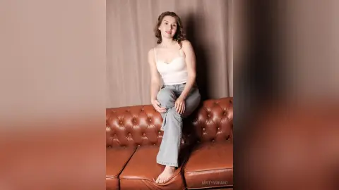 Smiling young woman in a white camisole and blue jeans sitting on a brown leather sofa in a bright room.