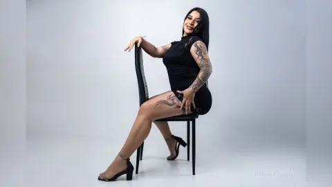 BellaMonrow poses sitting sideways on a black chair, wearing a short black dress and black high-heeled sandals, showing off her arm and leg tattoos.