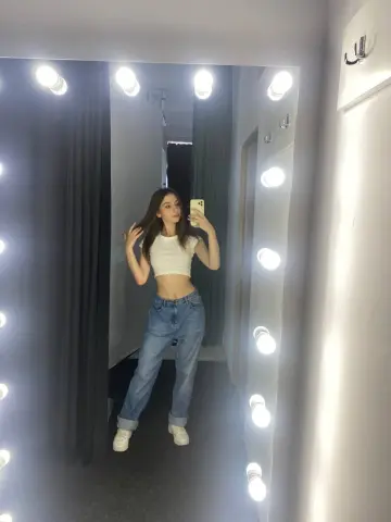 Young woman in white crop top and blue jeans taking a selfie in a bright lighted dressing room mirror.