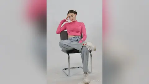 Young woman in pink turtleneck and light wash jeans sitting on a chair in a minimalist studio setting.