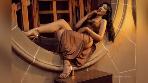 Elegant woman in a bronze satin dress lounging in a sunlit stone alcove with a warm, glamorous mood.