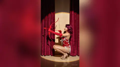 Elegant woman in red bodysuit and pearl details posing with a bow against velvet curtains in dramatic lighting.