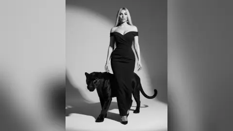 Kym stands in a long, off-the-shoulder black dress against a plain background.