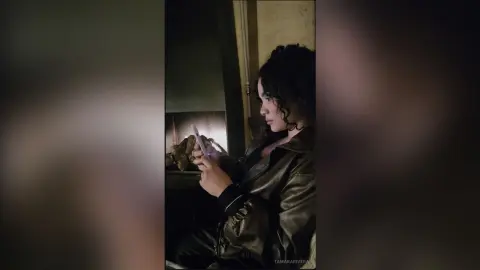 Young woman with curly hair in a black leather jacket using a smartphone in a moody, dimly lit indoor setting.