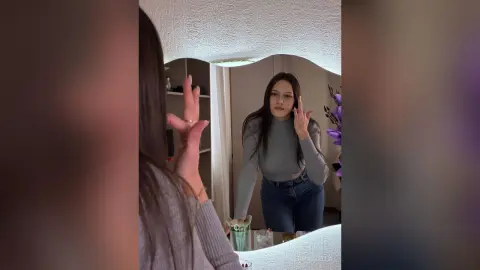 IngerMarzella poses in a grey long-sleeve top and blue jeans, looking into a mirror with one hand raised near her face.