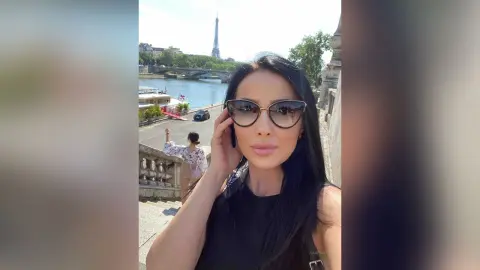 Stylish woman in black sunglasses and top smiling near the Eiffel Tower in Paris on a sunny day.