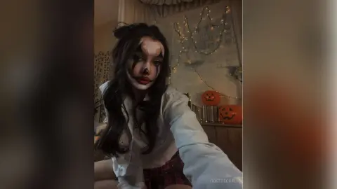 Woman in white blouse and plaid skirt with spooky clown makeup sitting near Halloween pumpkins.