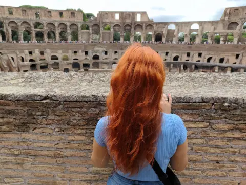 LeilaBlissXO shows her long red hair from behind while wearing a blue t-shirt, standing in front of the stone arches of the Colosseum.
