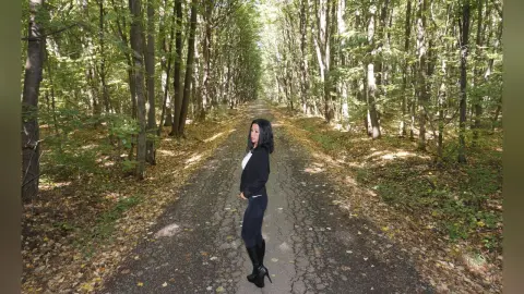 Young woman in black outfit standing on a sunlit forest path surrounded by lush green trees.
