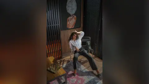 Stylish woman in white blouse and black trousers posing confidently in a modern, moody interior setting.