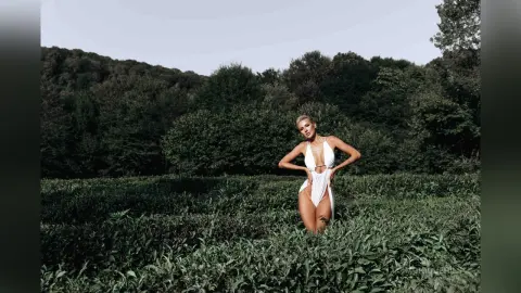 Elegant woman in a white bodysuit posing in a lush green forest field under a soft sky.