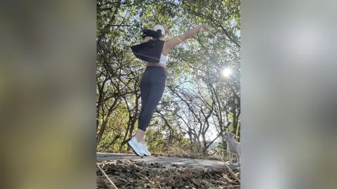 Woman in black leggings and white crop top stretching outdoors in a sunny wooded park with her dog.
