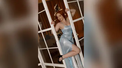 Elegant redhead woman in a shimmering blue evening gown posing by a window near a festive Christmas tree.