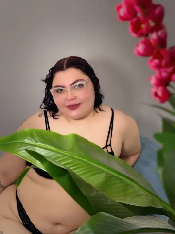 Smiling woman in black bikini relaxing behind large green tropical leaves in a bright indoor setting.