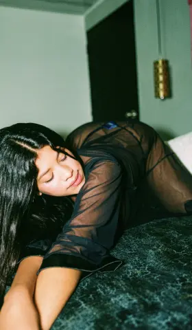 Young woman in a black sheer mesh outfit relaxing on a bed in a moody, dimly lit bedroom setting.
